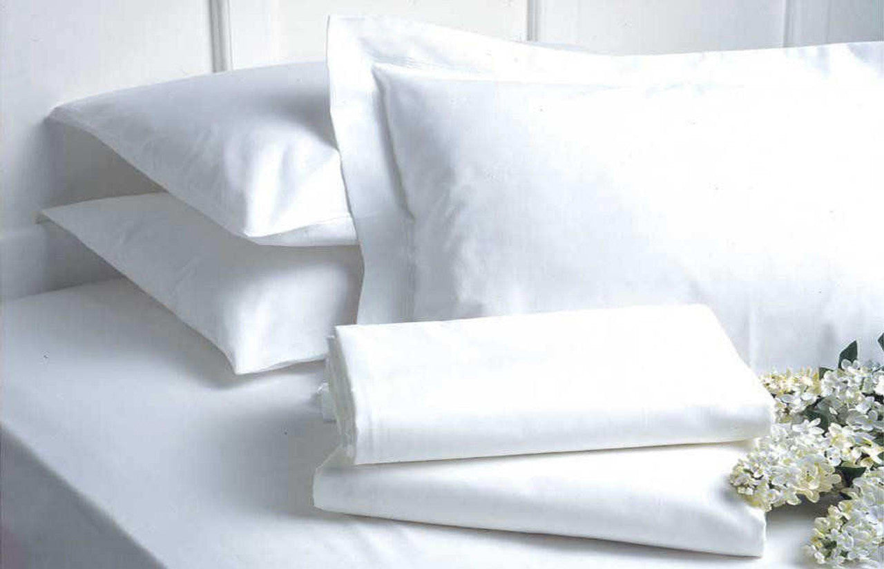 A neatly made bed with Ganesh Mills’ Oxford Micro Superblend Bed Linen fitted sheets and pillows, a folded white sheet on top, and a small bunch of white flowers placed beside it.