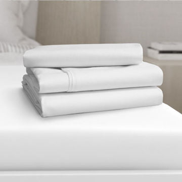 A neatly folded stack of Ganesh Mills Flat Sheets 2" Top Hem & 1" Bottom Hem - Oxford Micro Superblend Bed Linen rests on a white mattress, with a softly blurred bedroom in the background.
