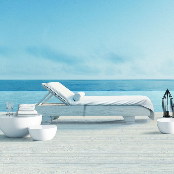 A Ganesh Mills Oxford Lounge Cover Towel in White/Beige drapes a white lounge chair on a light wood deck by the calm blue sea, accented with minimalist white décor and a black lantern under clear skies.