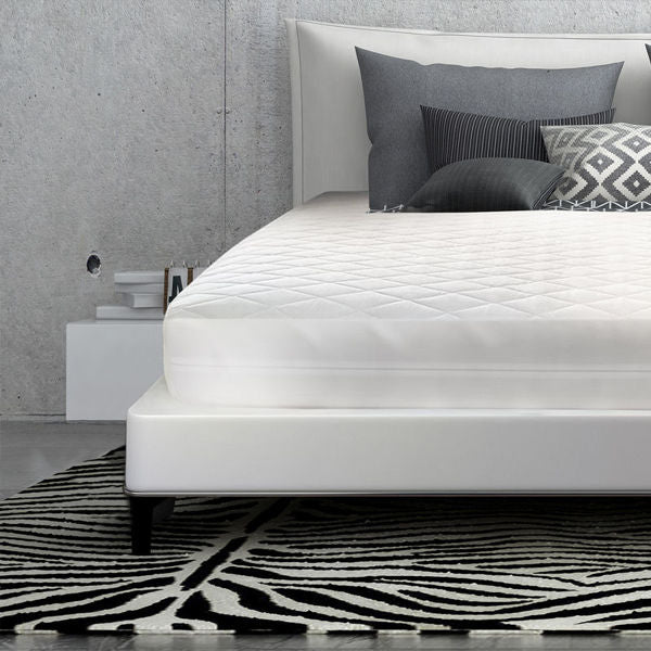 A modern bedroom with a Ganesh Mills Oxford Bed Bug Quilted Mattress Encasement with built-in pad on a low frame, geometric black and white rug, concrete walls, two pillows, and a headboard with stacked books on the side table.