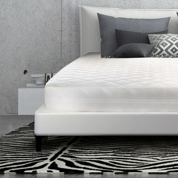 A modern bedroom with a white upholstered bed, Ganesh Mills 15 Pocket - Oxford Bed Bug Protective Luxurious Cover with Zipper & Velcro, gray and patterned pillows, a black-and-white geometric rug on concrete, and a white nightstand against a gray wall.