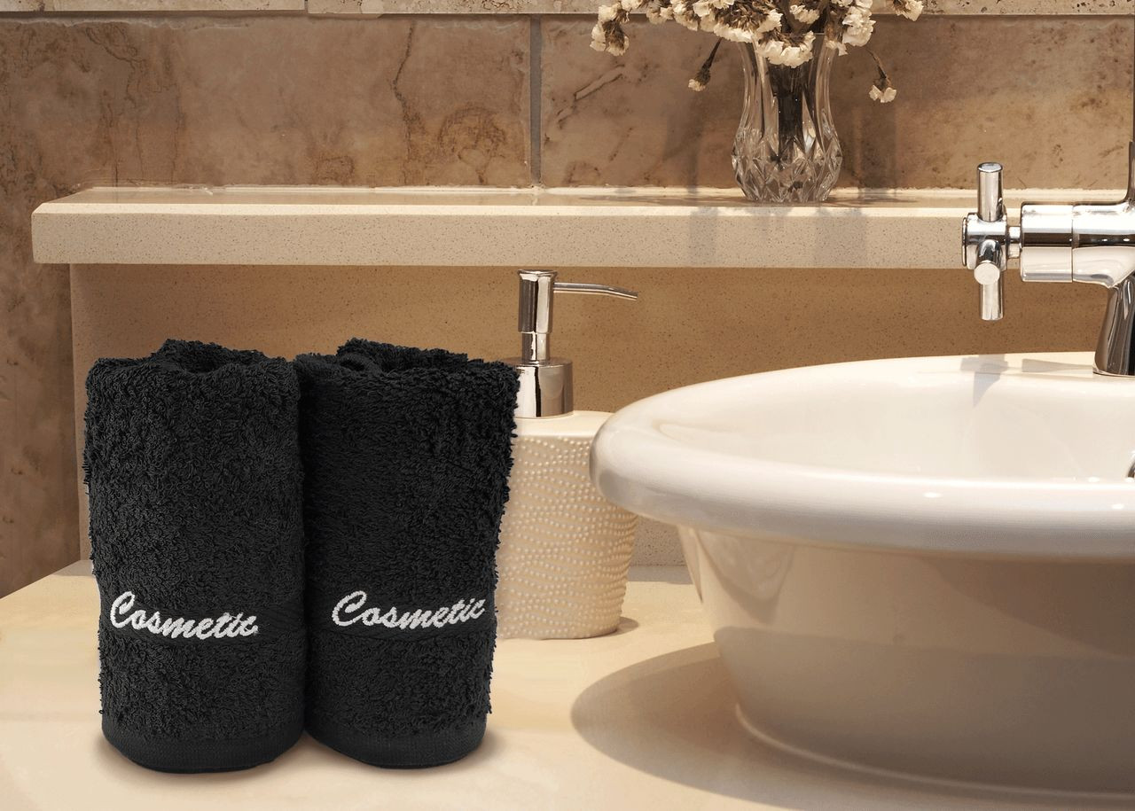 Two black "Cosmetic" embroidered cotton washcloths by Ganesh Mills rest on a bathroom counter near a white soap dispenser and sink, complemented by beige tiles and dried flowers—perfect for adding style to your daily routine.