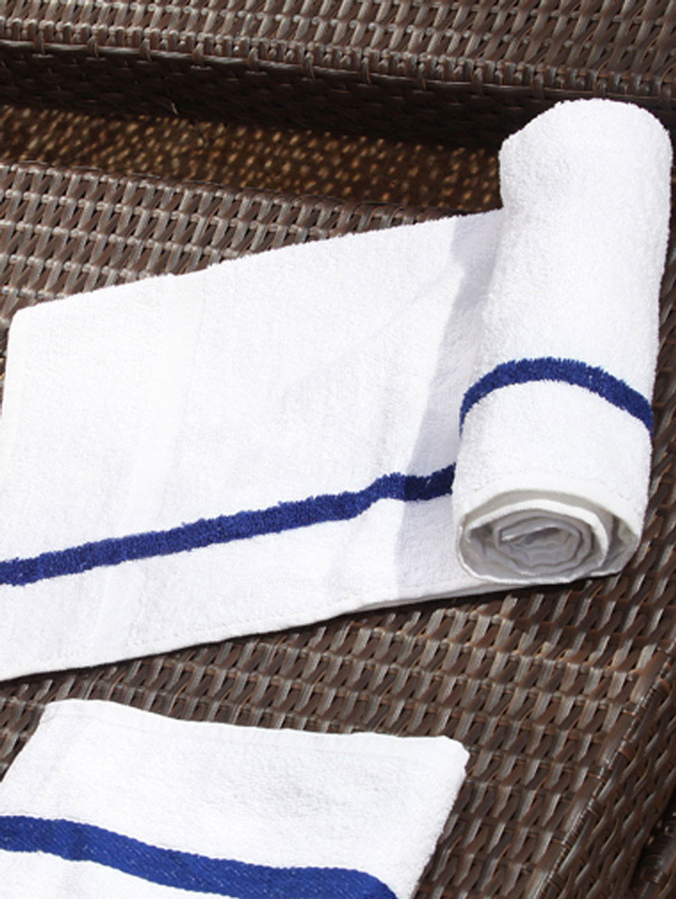 A Ganesh Mills White w/ Blue Center Stripe Pool Towel - Oxford Wizard is partially rolled on a brown woven surface, next to an unrolled towel of the same style.