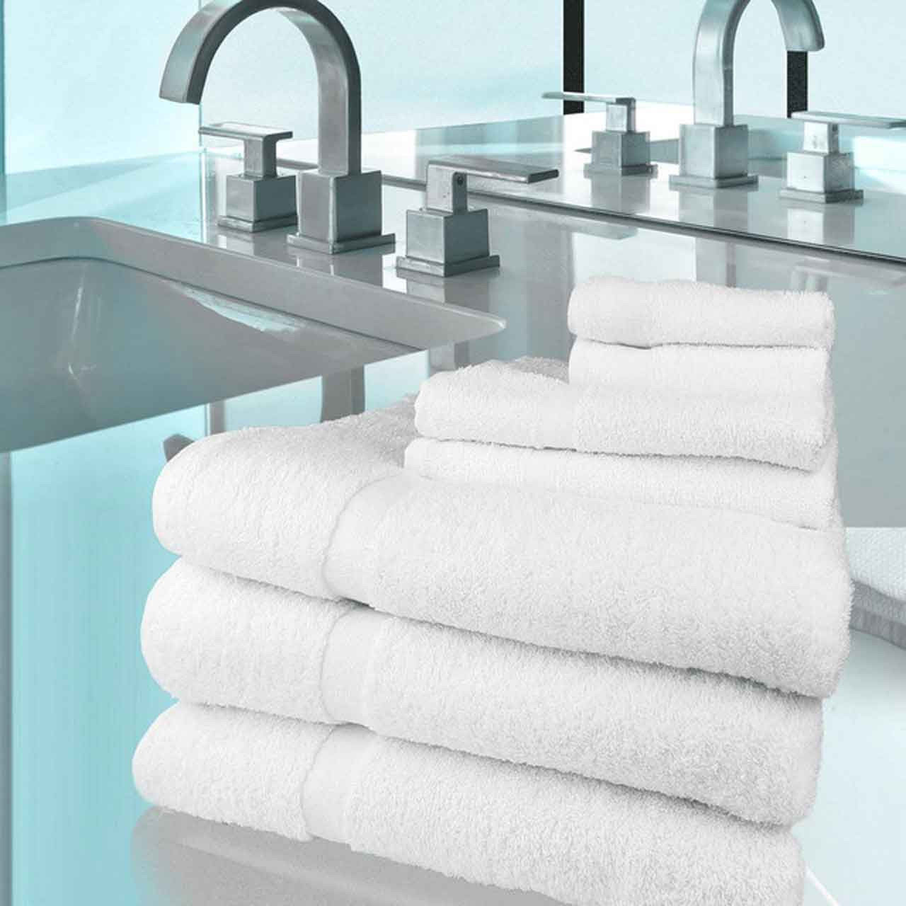 A stack of neatly folded Ganesh Mills Washcloth Hemmed - White Oxford Gold Cam Towels rests on a countertop in a modern bathroom featuring a sink, shiny silver faucets, and a clean, minimalist design.