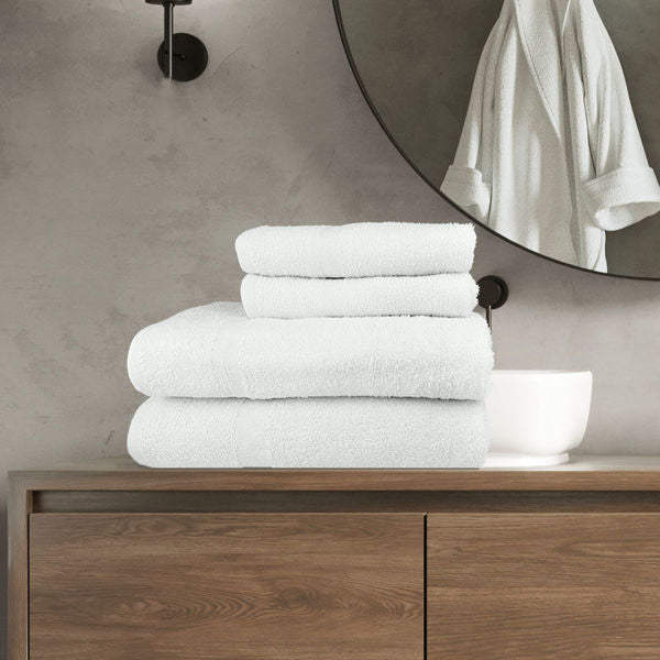A stack of Ganesh Mills white Oxford Classic Bronze towels sits on a wooden cabinet above a plush matching bathmat, with a round mirror reflecting a white bathrobe, all set against a gray concrete wall and black light fixture.
