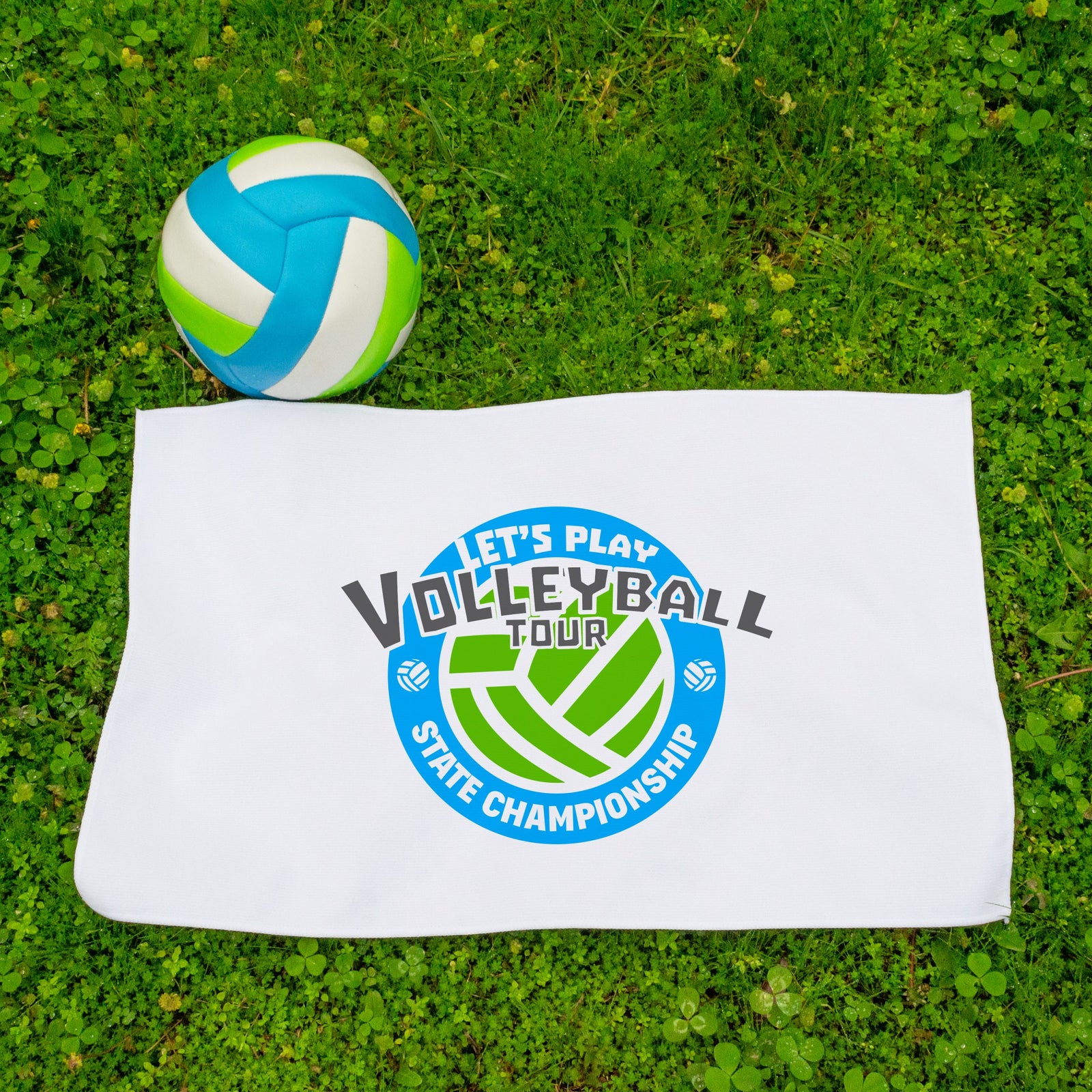 A blue, green, and white volleyball sits on grass next to a Pro Towels Microfiber Sports Towel featuring a Let’s Play Volleyball Tour State Championship logo with a dynamic volleyball graphic.