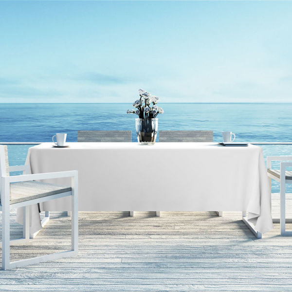 A table with a Ganesh Mills Oxford Banquet Table Linen in multicolor and two chairs sits on a deck over a calm ocean. A vase with white flowers and two cups rest on the table beneath a clear blue sky, creating a tranquil seaside atmosphere.