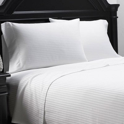 A neatly made bed with white striped sheets and two matching pillows, set against a dark, ornate wooden headboard, features the Millennium - Duvet Cover Stripe by WestPoint Home—an elegant, hotel-quality duvet cover crafted from premium commercial linen.