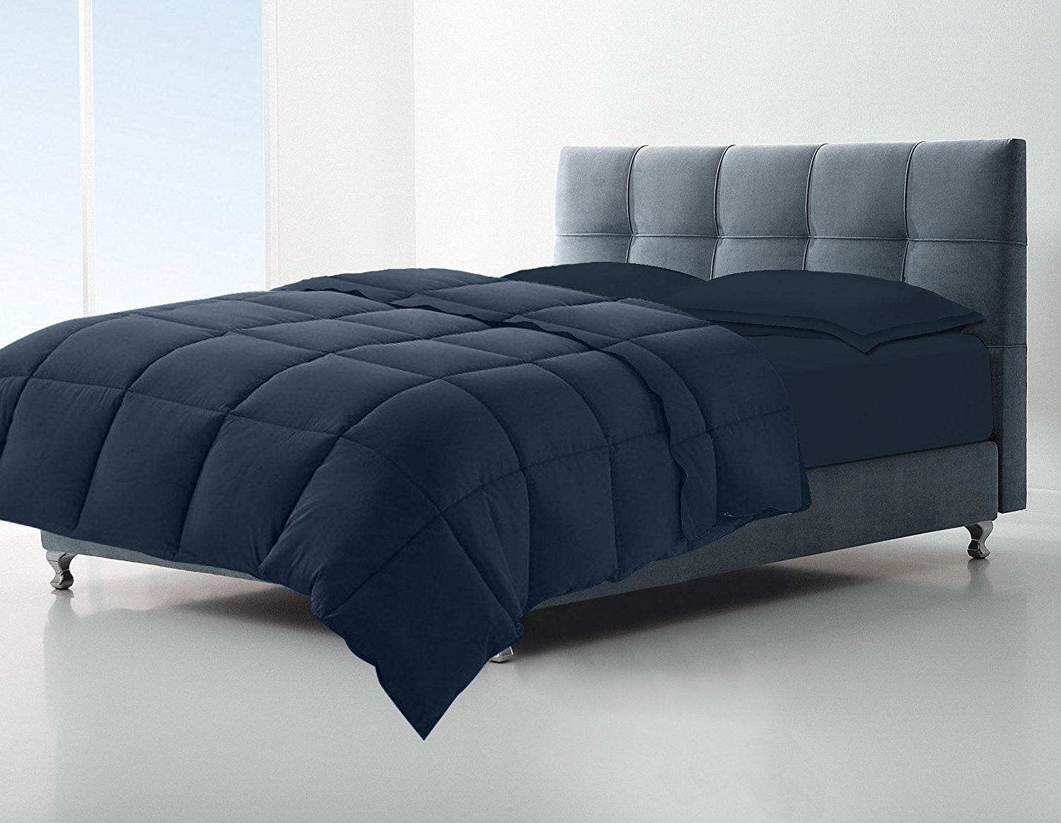 A modern bed with a tufted headboard is styled with the KSE Down Alternative Comforter in navy blue and matching pillows, set in a bright, minimal room with white walls and large windows.