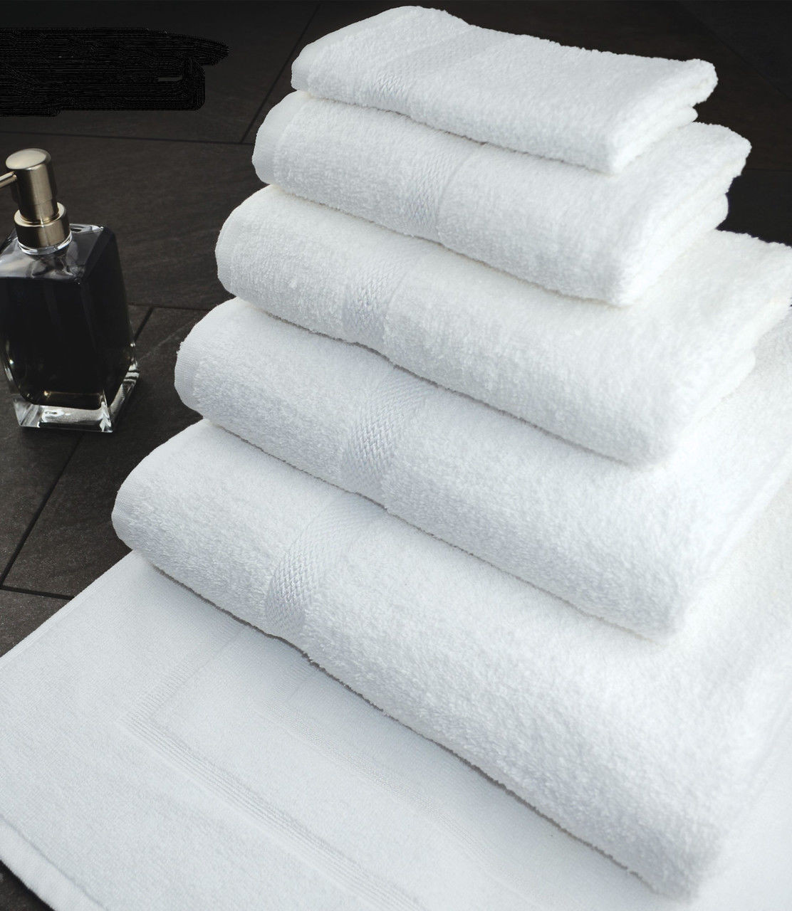 A set of Ganesh Mills Hand Towel - White Oxford Nuvola Towels, crafted from soft ringspun cotton in various sizes, is neatly arranged on a dark surface beside a square bottle with a silver pump.