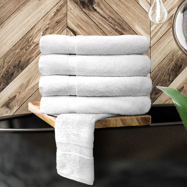 A stack of five Ganesh Mills Bath Sheet / Pool Towels - White Oxford Bellezza, crafted from soft cotton, is neatly folded on a wooden shelf with one towel draped over the edge. A chevron-patterned wall and a green plant add to the tranquil scene.