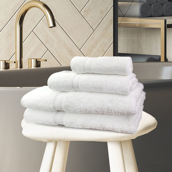 A neatly folded stack of Ganesh Mills Hand Towel - White Oxford Forza Towels rests on a stool in a modern bathroom featuring a gold faucet, freestanding tub, and herringbone-patterned wall tiles.