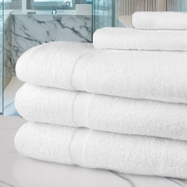 A stack of fluffy white Ganesh Mills Bath Towels from the Oxford Gold Cam Towel Collection is neatly folded on a marble countertop in a modern bathroom with large windows and marble walls.