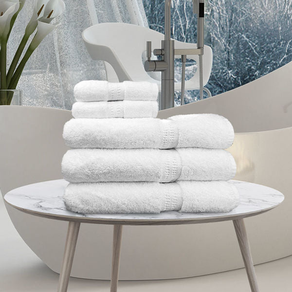 A stack of Ganesh Mills Bath Sheet / Pool Towels from the White Oxford Reserve Towel collection is arranged on a round white marble table in a modern bathroom featuring a freestanding tub, white chair, and vase of white flowers.