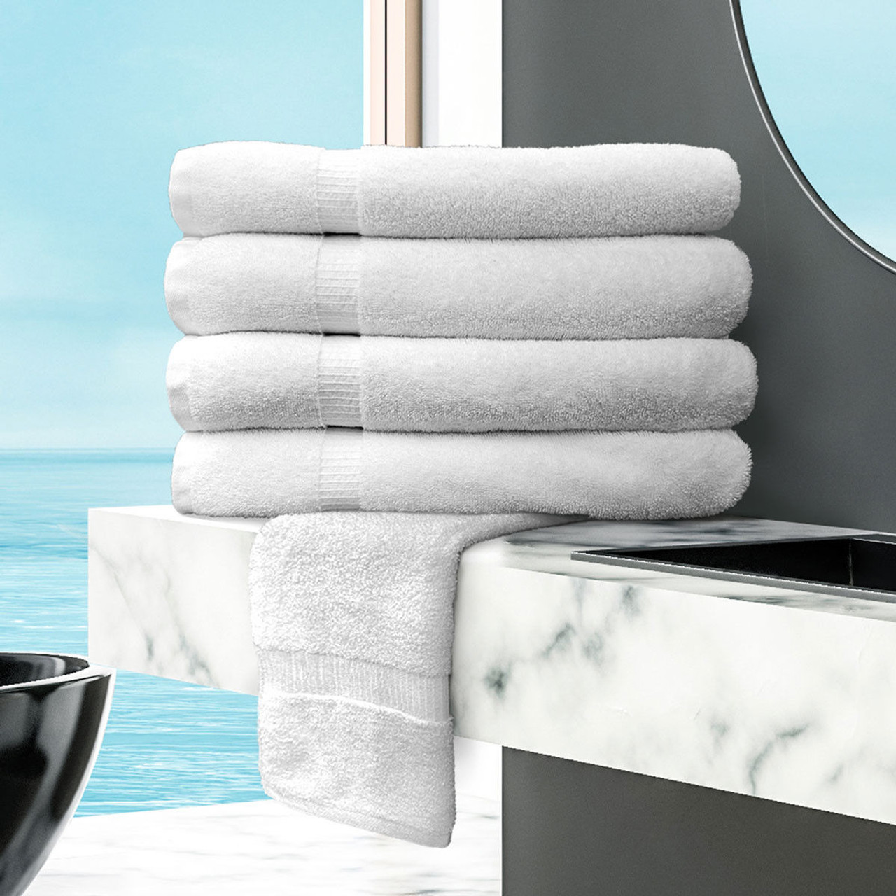 Five neatly folded Ganesh Mills Washcloth - White Oxford Bellezza Towels are stacked on a white marble countertop in a modern bathroom with a large round mirror and an ocean view visible through the window.