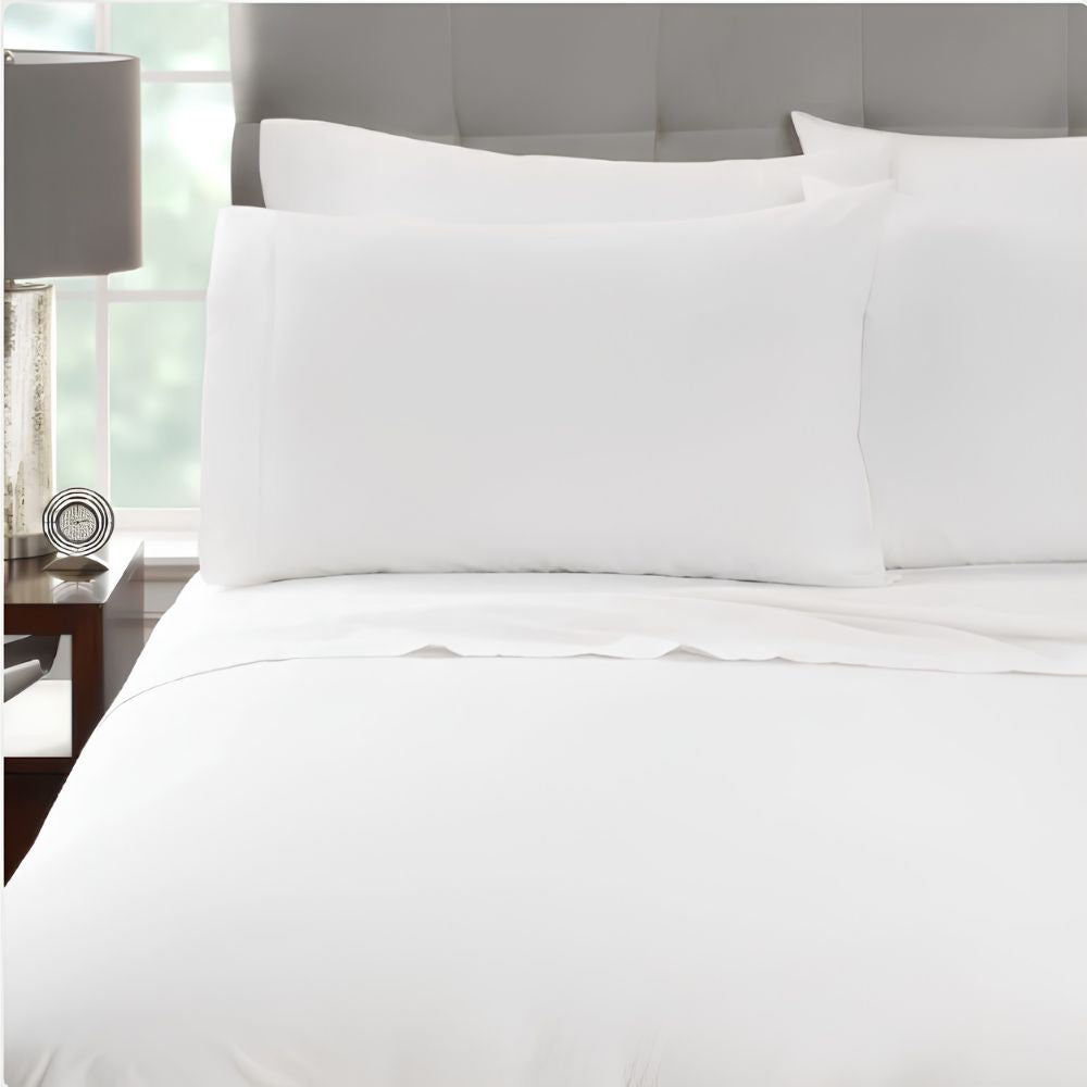 A neatly made bed with WestPoint Home’s Martex Millennium 300 T300 Percale Sheets & Pillowcases sits by a nightstand with a lamp, featuring a tufted gray headboard and a window in the background of a bright room.