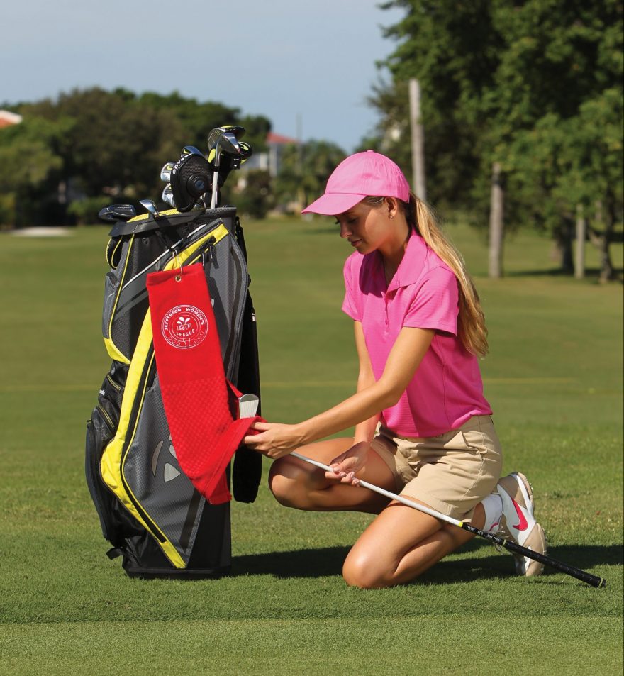 A woman in a pink cap and shirt kneels on a golf course beside her bag, holding Pro Towels’ “Scrubber” Towel with custom embroidery. Trees and blue sky are visible in the background.