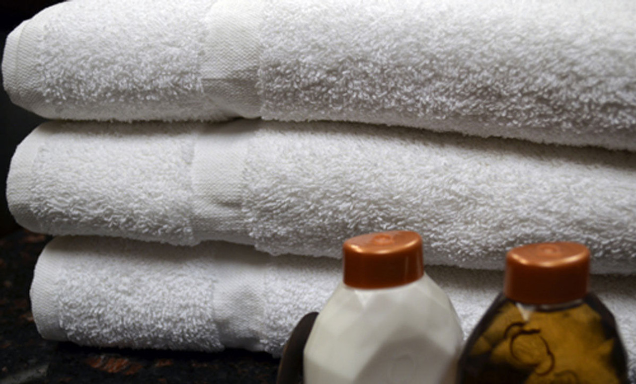 Three neatly folded white washcloths from the Ganesh Mills Oxford Silver Towel collection are stacked on a dark surface. In front, two small bottles with brown caps—likely hotel toiletries—are displayed.