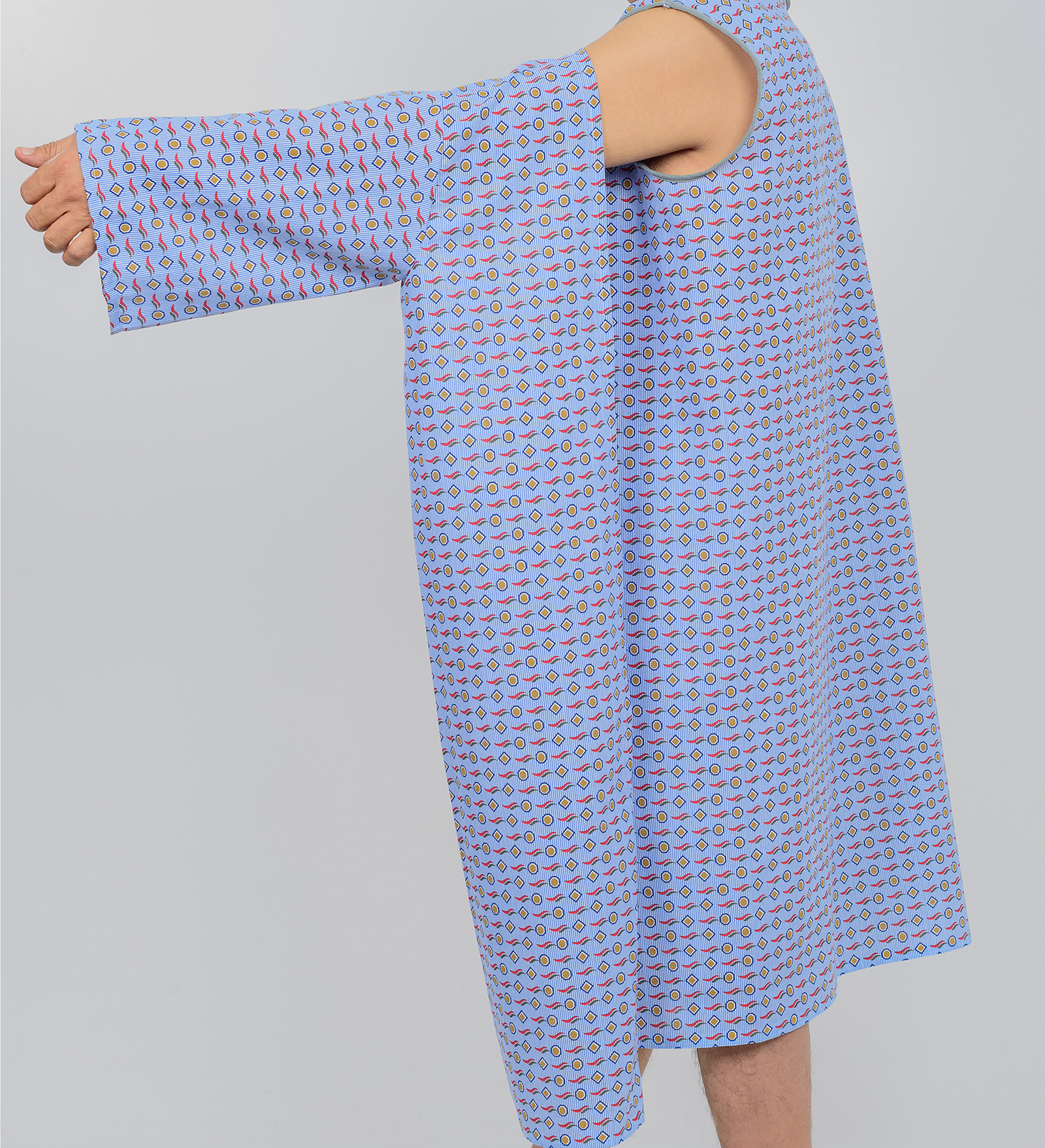 A person wears a KSE Three-Arm hole Gown, a loose-fitting hospital gown with a blue and pink pattern, extending their right arm to the side against a plain light background. The oversized gown provides full body coverage.