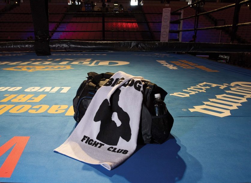 A gym bag, a Pro Towels Jewel Collection Sport Towel in white, and a water bottle rest on the edge of a blue boxing ring, with empty arena seats visible in the dim background.