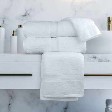 A stack of Ganesh Mills Bath Towel - White/Ivory Oxford Vicenza Towels rests on a white marble bathroom counter with gold handles, accompanied by white toiletry containers.