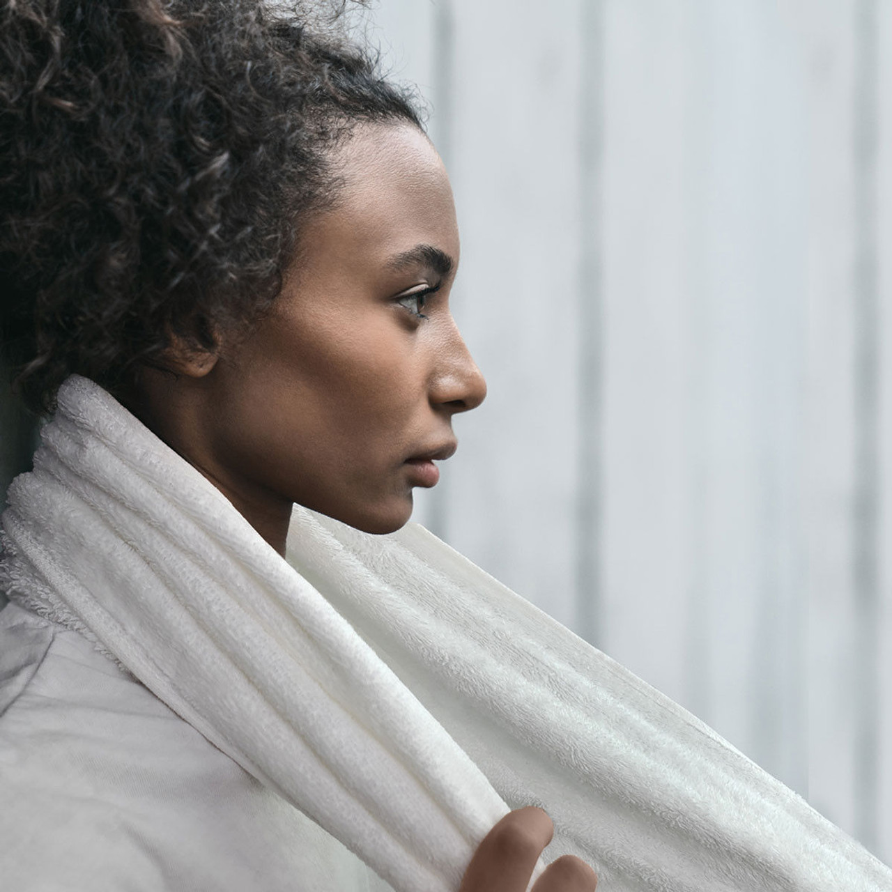 A young woman with curly hair in a ponytail stands in profile, wearing a white top and the Ganesh Mills Sports Neck Towel Hemmed - White Velour Towels draped around her neck, set against a light gray background.