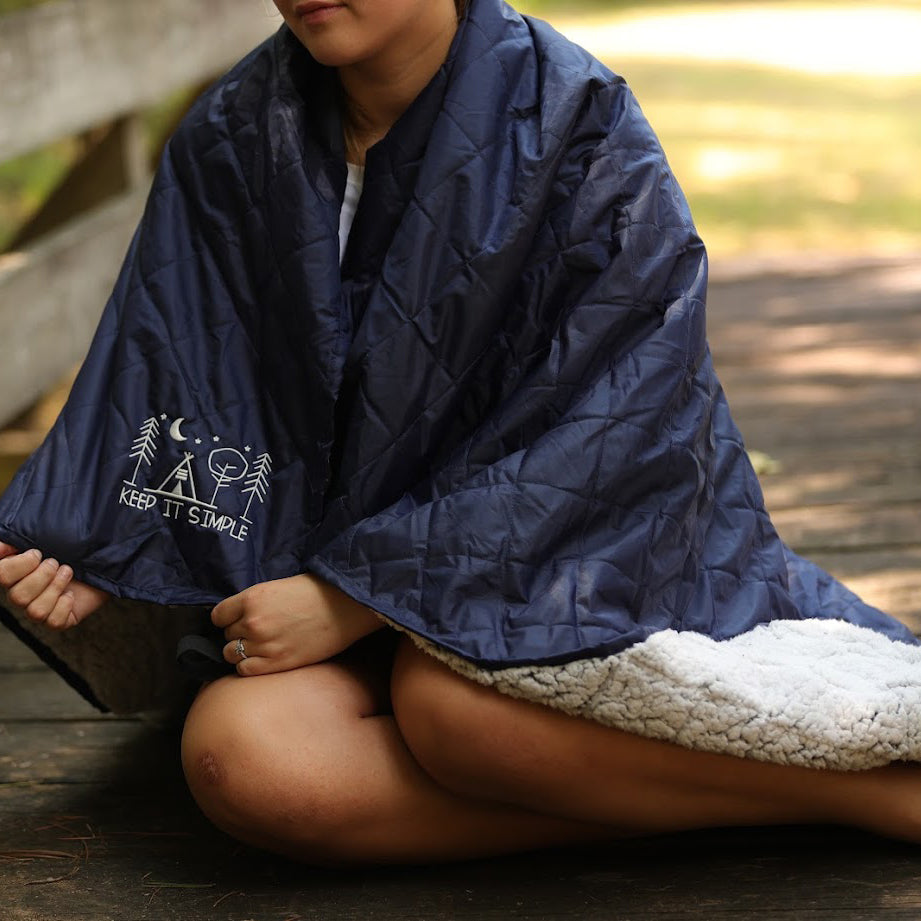 A person sits cross-legged outdoors, wrapped in the Pro Towels Waterproof Sherpa Blanket, featuring a simple camping design and "KEEP IT SIMPLE" text—ideal for cozy moments or custom embroidery.