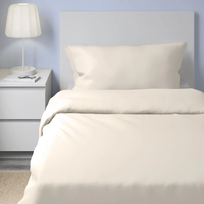 A single bed features cream-colored organic cotton sheets and pillowcase, plus the Bargoose AllergyCare Organic Cotton Zippered Duvet Encasement, beside a white nightstand with a lamp, glasses, and book, all against a light blue wall.