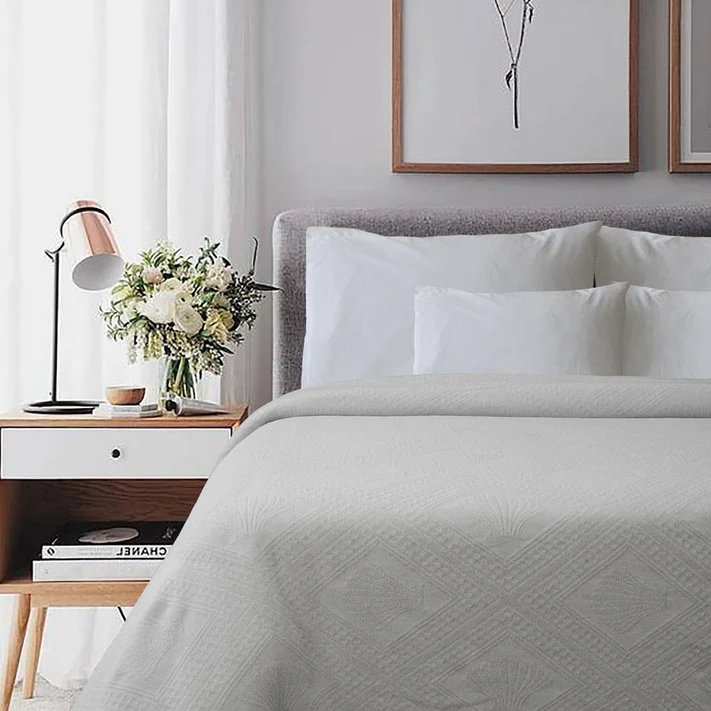 A neatly made bed features the Avalon Jacquard Bedspread by Bargoose, paired with white pillows. Beside it stands a wooden nightstand with a flower vase, books, and lamp. Above the bed, two framed artworks complete this hotel-inspired look.