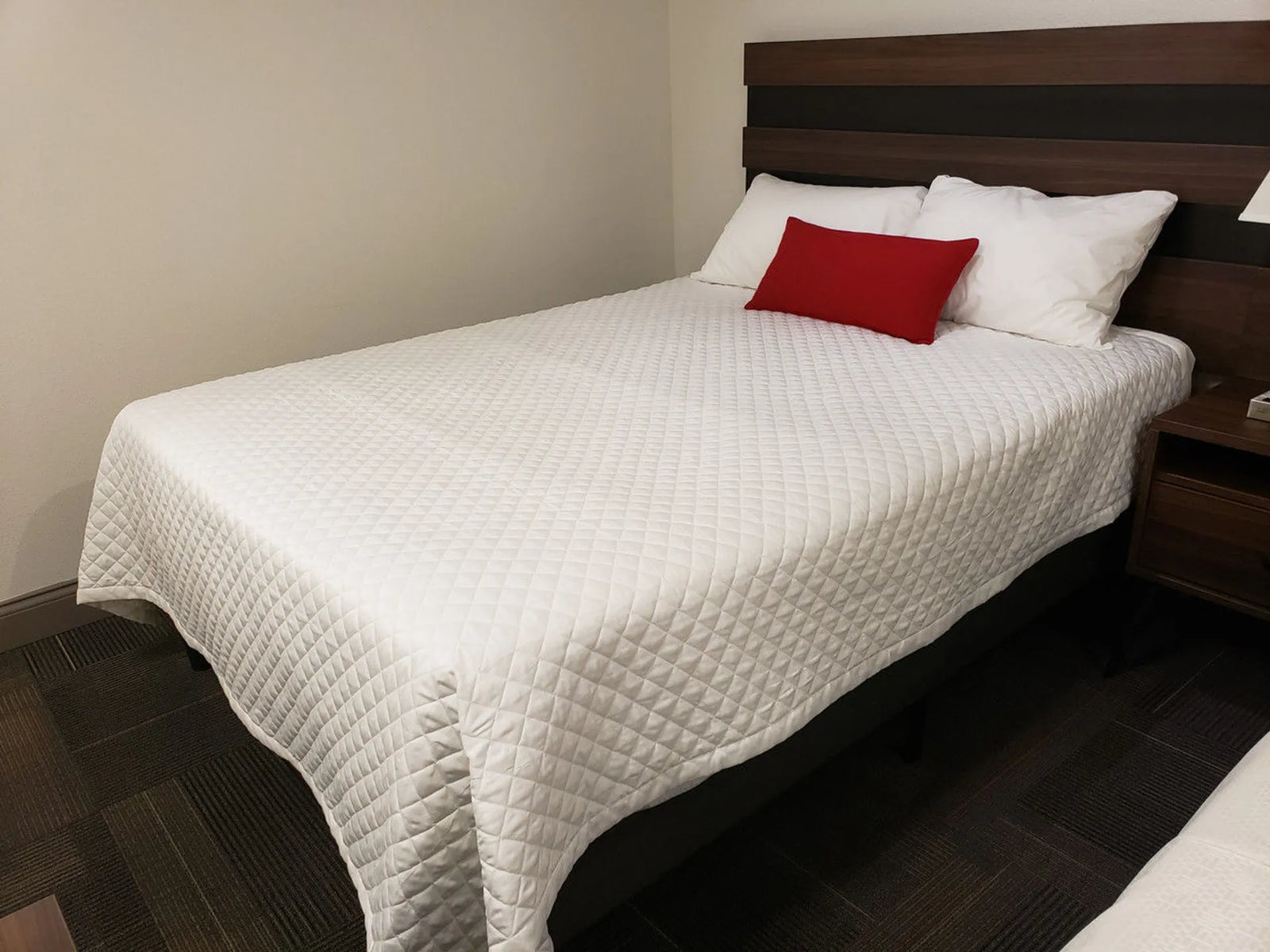 A neatly made bed features the Ganesh Mills Diamond Check Quilted Bed Topper - Oxford Ultra Light Duvet/Coverlet, three white pillows, a red accent pillow, and a dark wooden headboard in a clean, neutral-toned carpeted room.