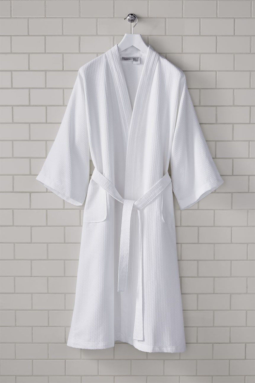 A white Diamond Sophi Shawl Robe by 1Concier with a lightweight diamond pattern and belt hangs on a hook against a light beige tiled wall.