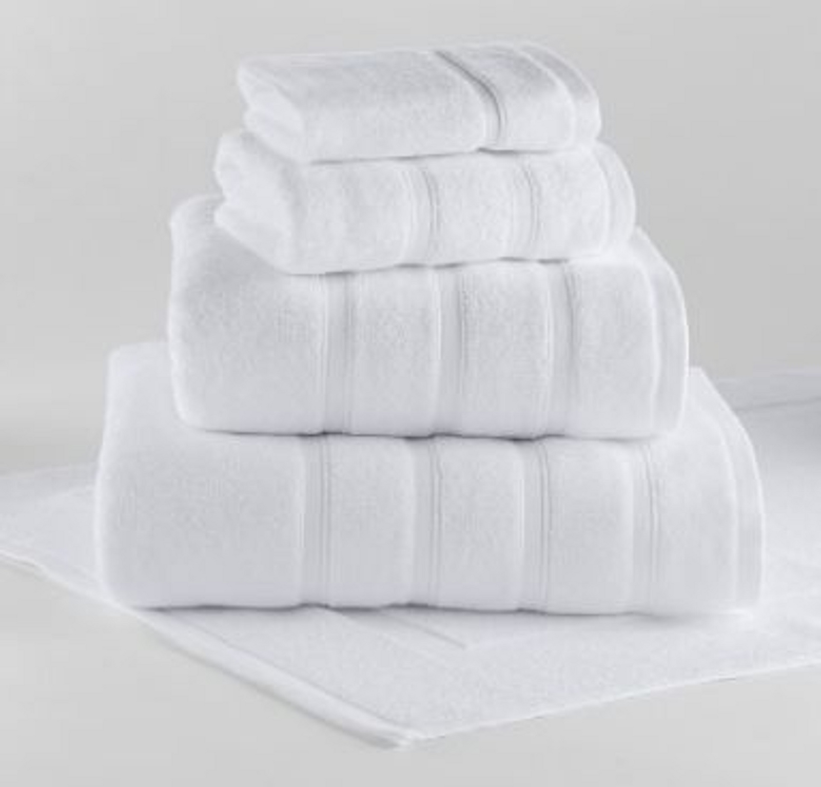 A neatly folded stack of white Bath Sheet Monaco Terry 100% Cotton towels by 1Concier in various sizes is arranged on a white surface, with one towel laid flat underneath, against a plain light background.