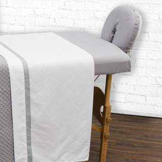 A massage table with a gray, stain-resistant cover and padded face cradle is topped with 1Concier Fitted Sheet Spa Sheets, standing on a wooden floor against a white brick wall.