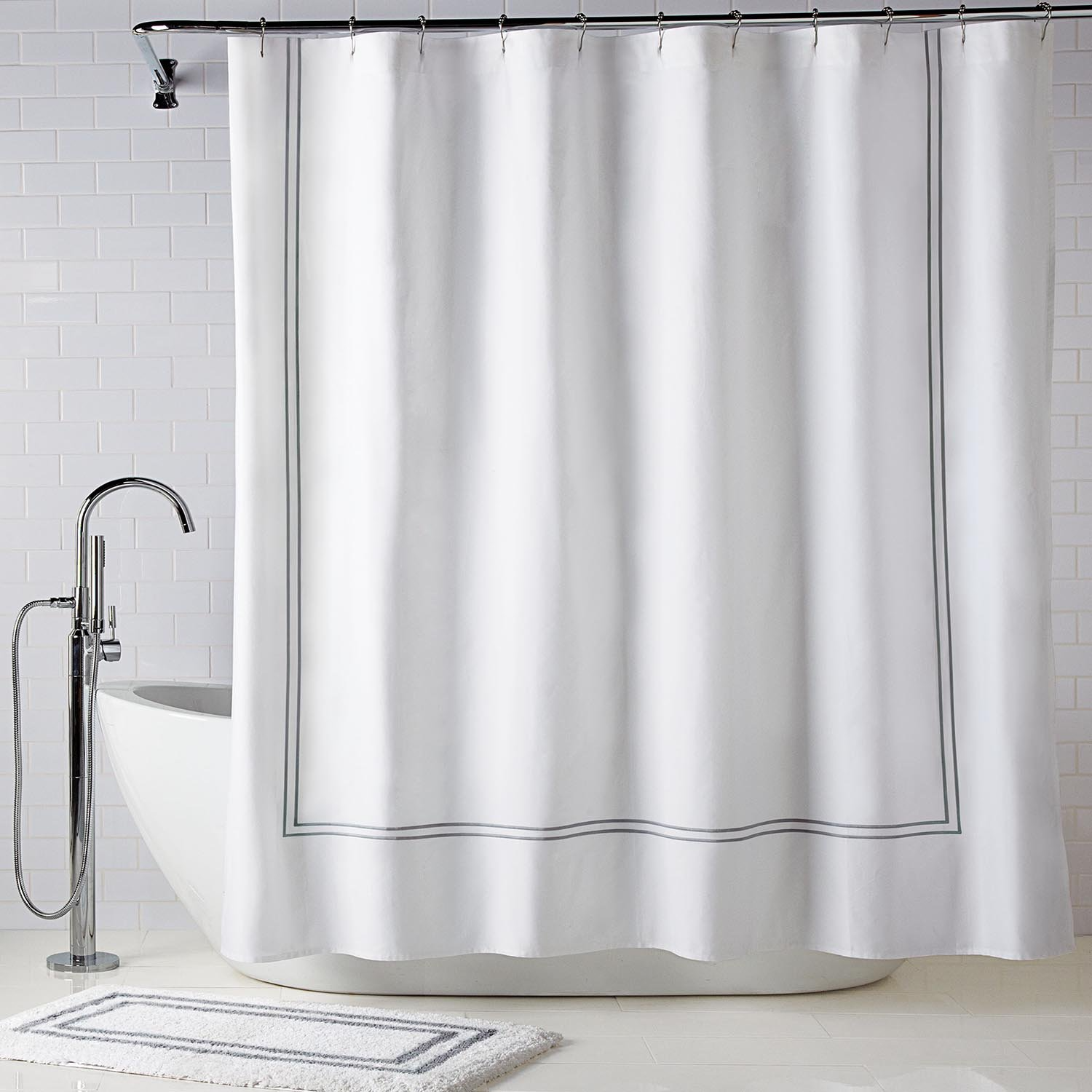 A white Supima cotton shower curtain with a thin gray border hangs above a freestanding tub. In front lies the Tufted Cut Bath Rug from the Gryphon Collection by WestPoint Home, on classic white subway tiles.