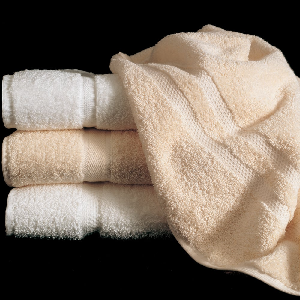 Three Martex Sovereign Dobby Towels by WestPoint Home—two white, one beige—are stacked on a black background, with a fourth beige towel draped loosely over the top.