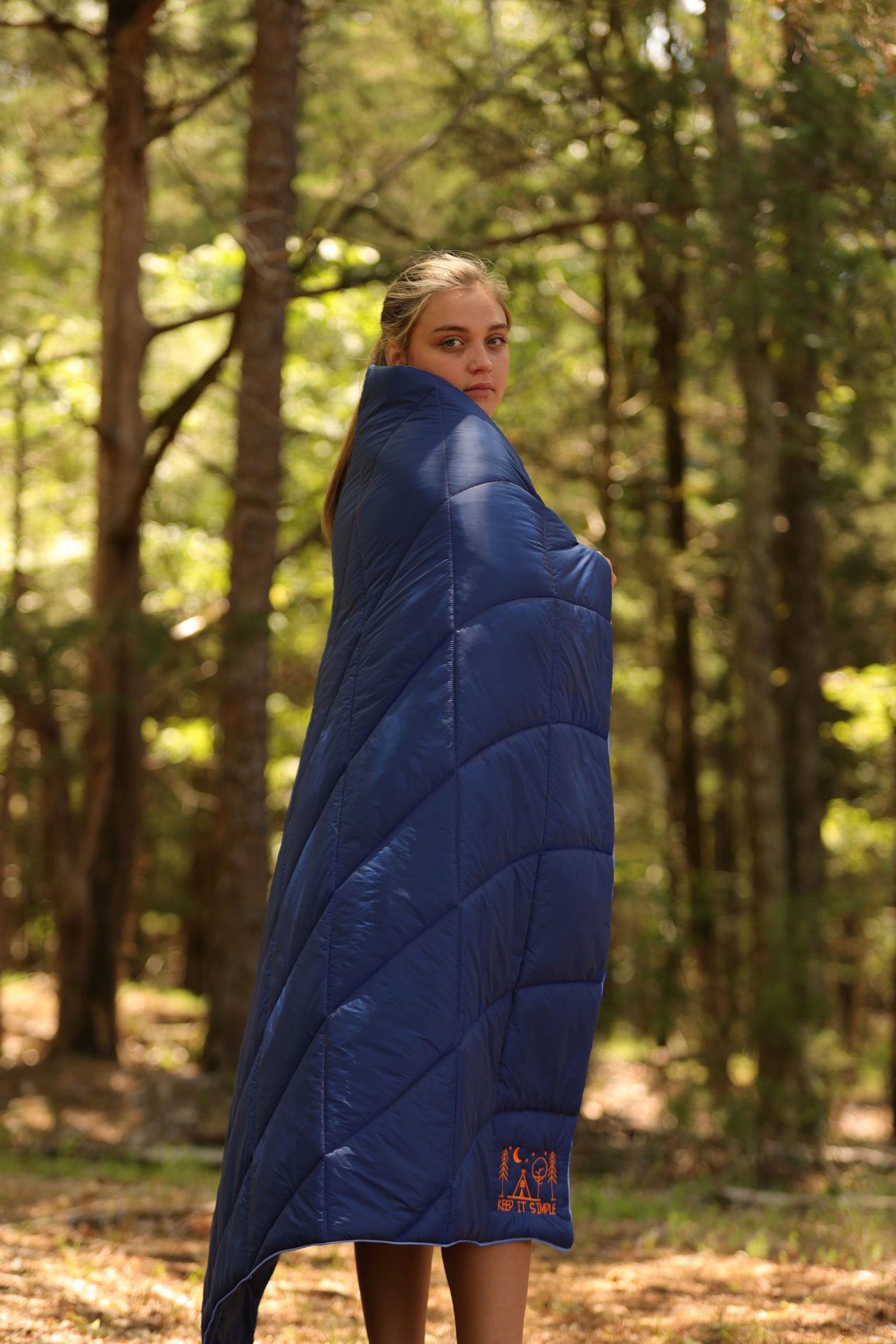 In a sunlit forest, a person wrapped in a Pro Towels Lofthouse Camping Blanket with orange embroidery glances over their shoulder at the camera, surrounded by trees and greenery.