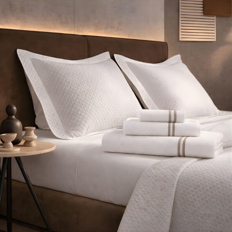 Luxury hotel bedroom display with white quilted pillows, neatly folded bath towels and premium white bedding on a modern upholstered bed.
