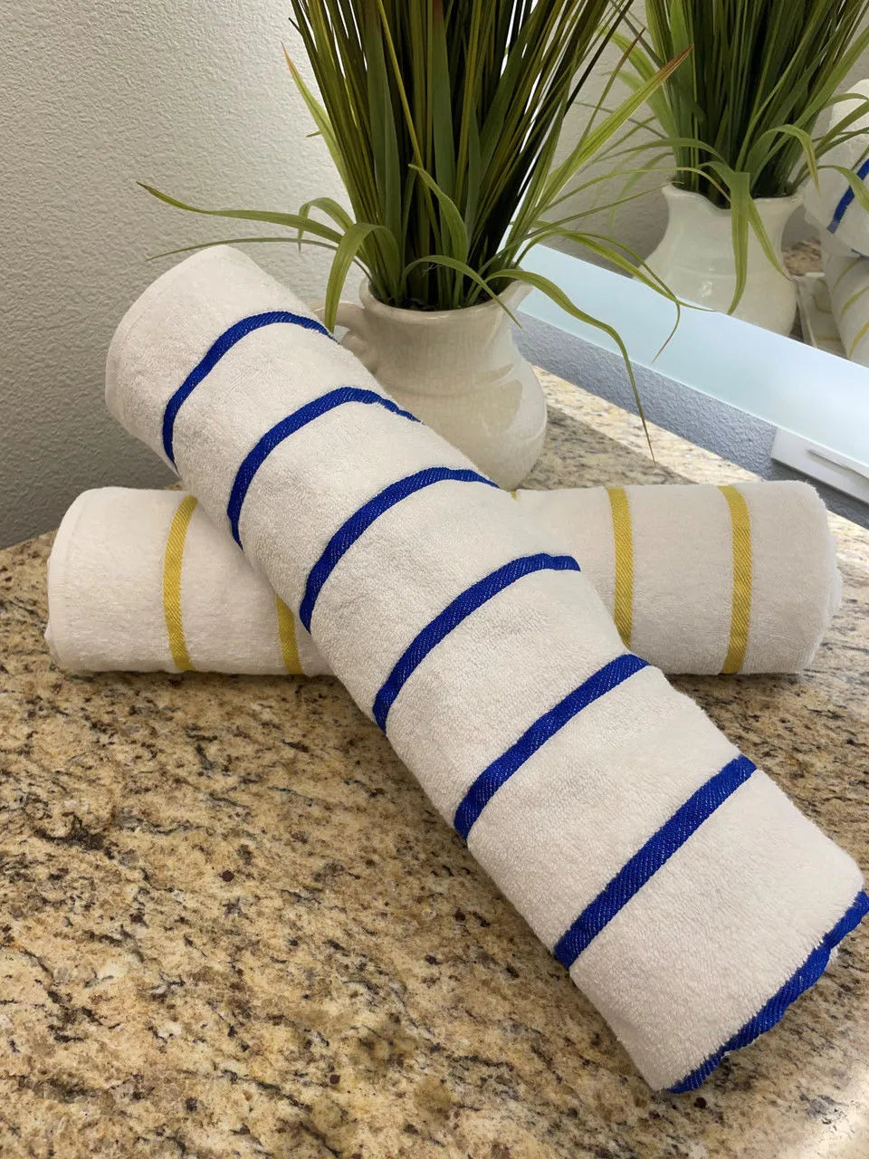 Two rolled Ganesh Mills Oxford Nautique Stripe Towels (white with blue/yellow stripes) rest on a granite countertop, with a white vase of greenery and a mirror behind—ideal for poolside towel displays.