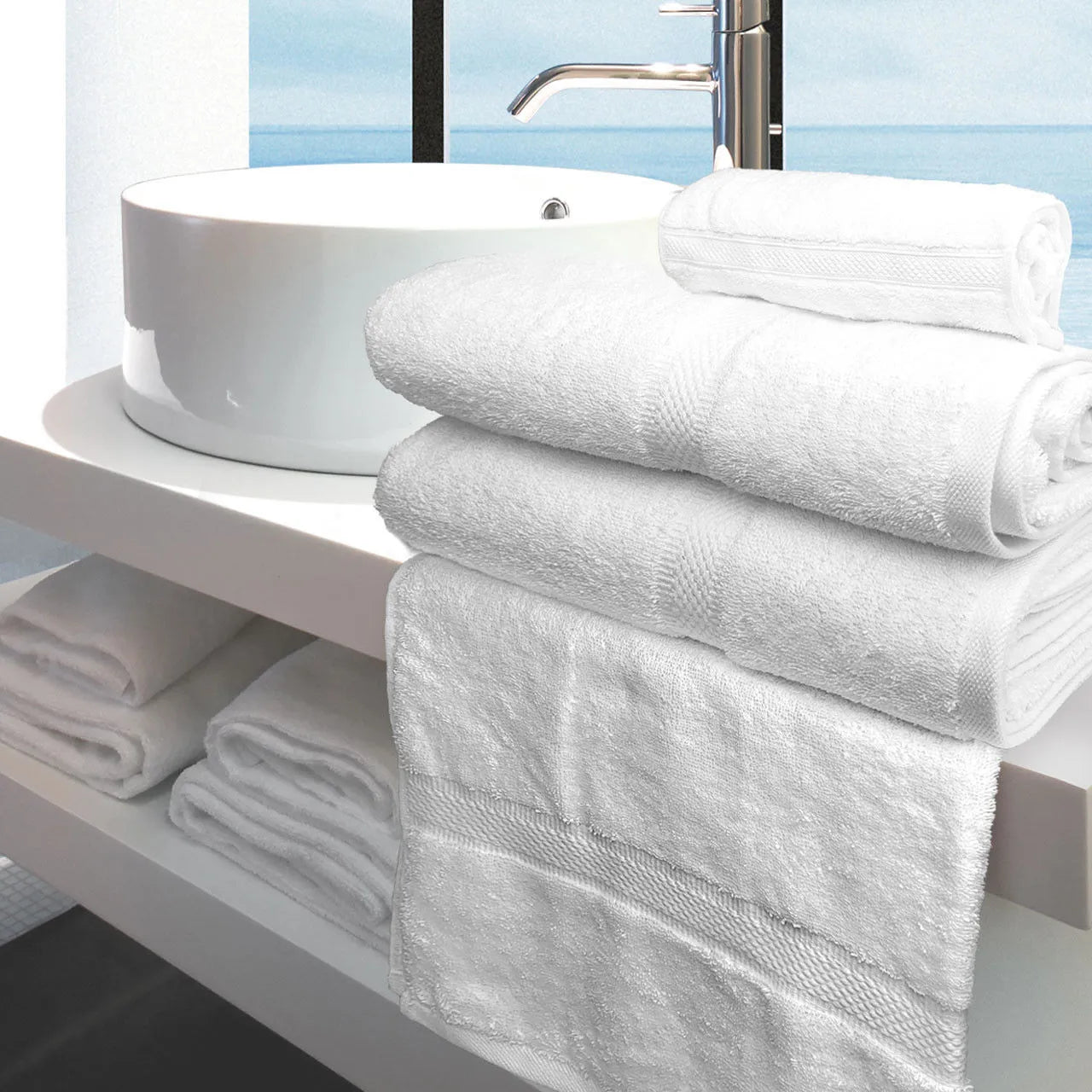 A stack of Ganesh Mills Washcloth - White Oxford Imperiale Max Towel Collection sits on a modern white bathroom counter with more towels and washcloths on a shelf below, and a window overlooking water in the background.