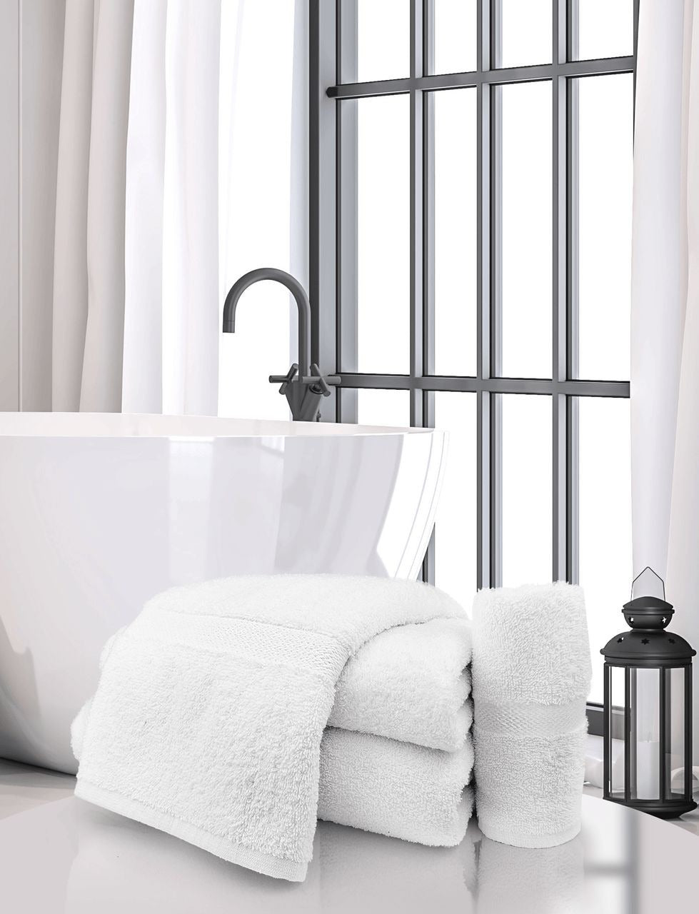 A bright, modern bathroom features a white freestanding tub, a stack of Ganesh Mills Washcloth - White Oxford Regale Towels, a rolled towel, a black lantern, and large windows with sheer white curtains letting in natural light.