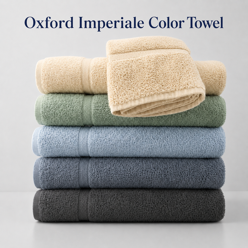 A stack of five Oxford Imperiale Color Towels by Ganesh Mills in beige, green, blue, light blue, and gray, plus a draped beige towel. Luxurious ringspun cotton—ideal for any bathroom.