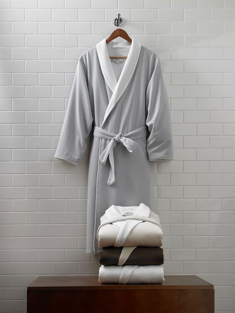 A light gray 1Concier Presidential Belt Robe with a white collar hangs on a wooden hanger against white tiles. Below, four luxurious 1Concier bathrobes in white, beige, light gray, and dark gray are stacked neatly on wood.