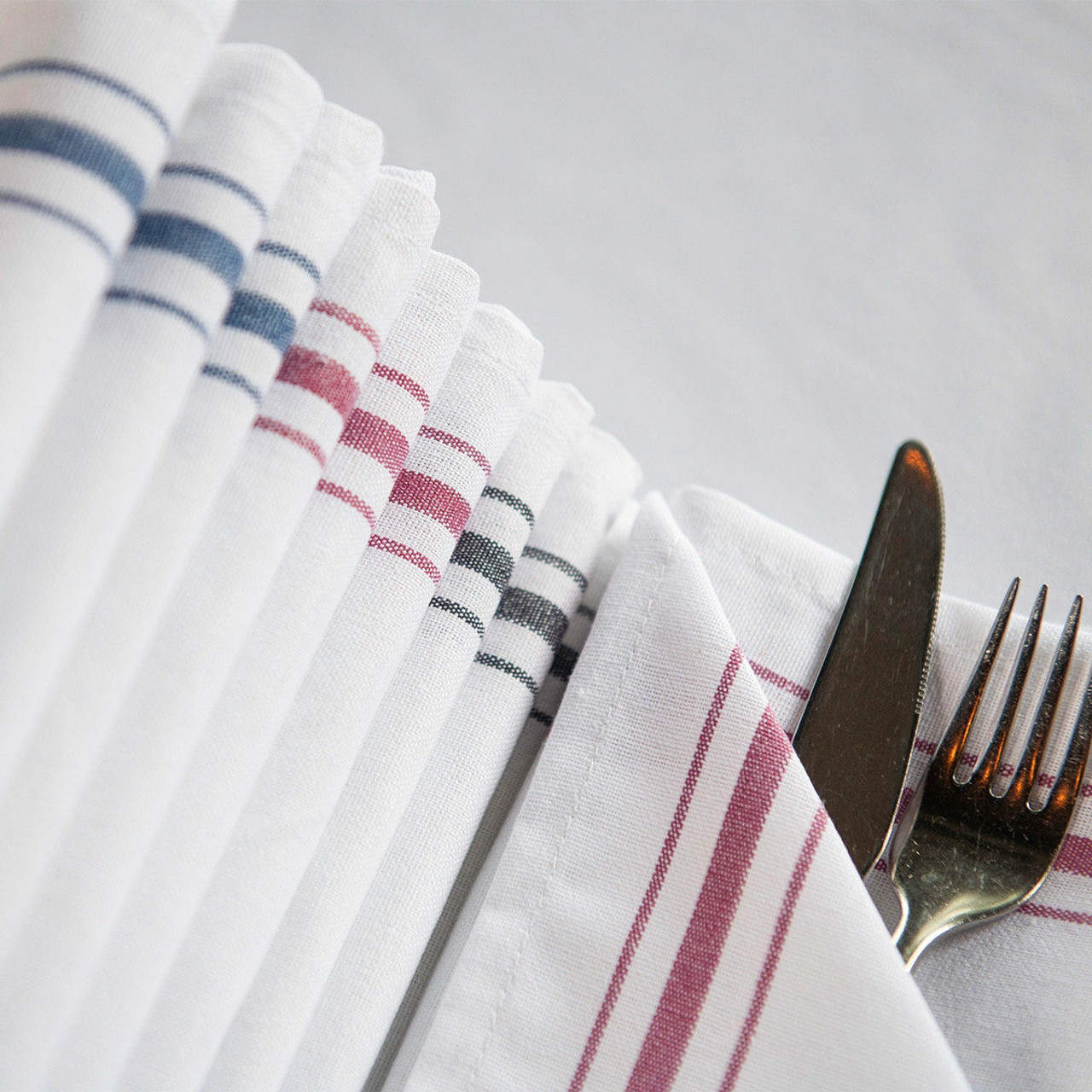 A close-up of neatly folded Napkin Bistro 100% Cotton White / Black Stripe by 1Concier, beside a knife and fork partially wrapped in one napkin, all set on a white tablecloth.