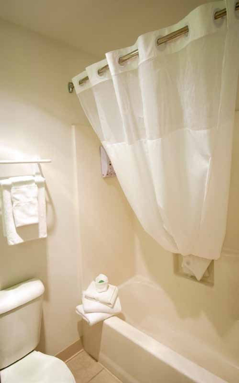 A small bathroom features a Ganesh Mills Hookless Shower Curtain with a 12" sheer voile window on the tub, a white toilet, a towel rack with white towels, and extra folded towels stacked neatly on the tub edge.