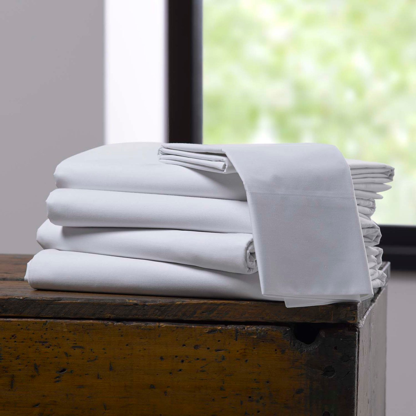 A neatly folded stack of Martex Green 250 eco-friendly bed sheets and pillowcases by WestPoint Home sits on a wooden surface near a sunlit window.