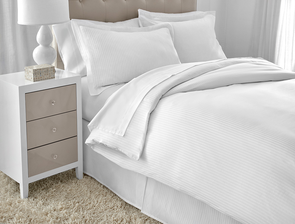 A neatly made bed with white striped bedding and pillows features the WestPoint Home Martex Ultra Touch FLEX Microfiber Down Alt Quilted Blanket. Next to it is a white three-drawer nightstand with a lamp and tissue box, set on a plush beige rug.