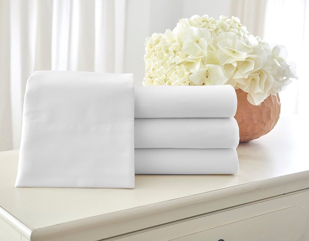 A set of WestPoint Home Five Star Hotel Collection DryFast 300 sheets and a pillow case in white is neatly stacked on a light dresser beside a vase of white hydrangeas, with sheer curtains in the background.