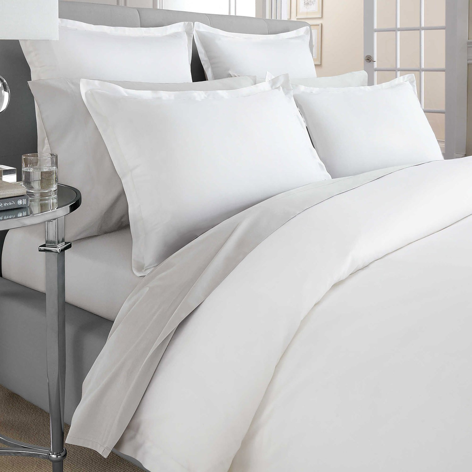 A neatly made bed features WestPoint Home’s Gryphon Collection Solid Duvet Cover in crisp white Supima cotton, with matching pillowcases and multiple upright pillows. A glass nightstand holds water and a book, creating a bright, hotel-style look.