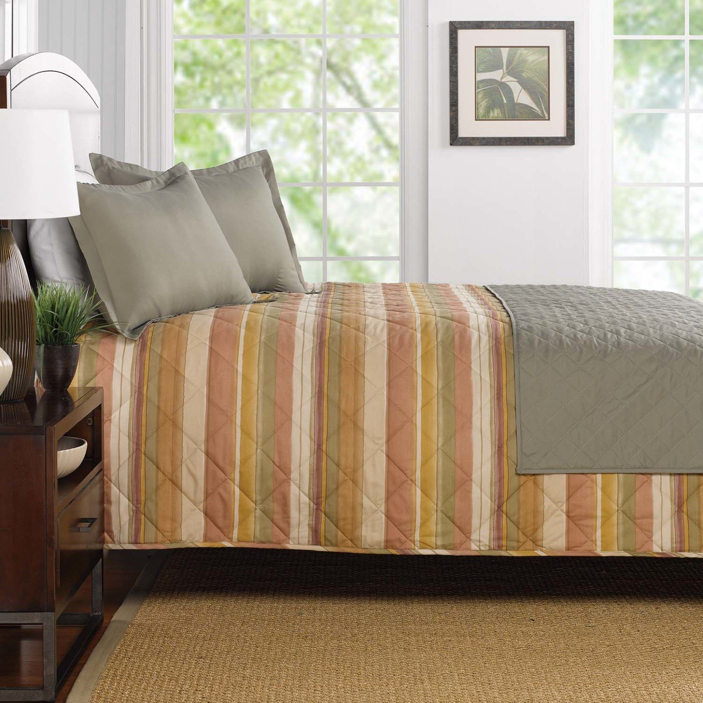 A neatly made bed features WestPoint Home's Coverlets - Westpoint Hospitality - Pattern in beige, yellow, and orange stripes, paired with two gray pillows and a gray quilt. The room includes large windows, a framed picture, and a nightstand with lamp and plant.