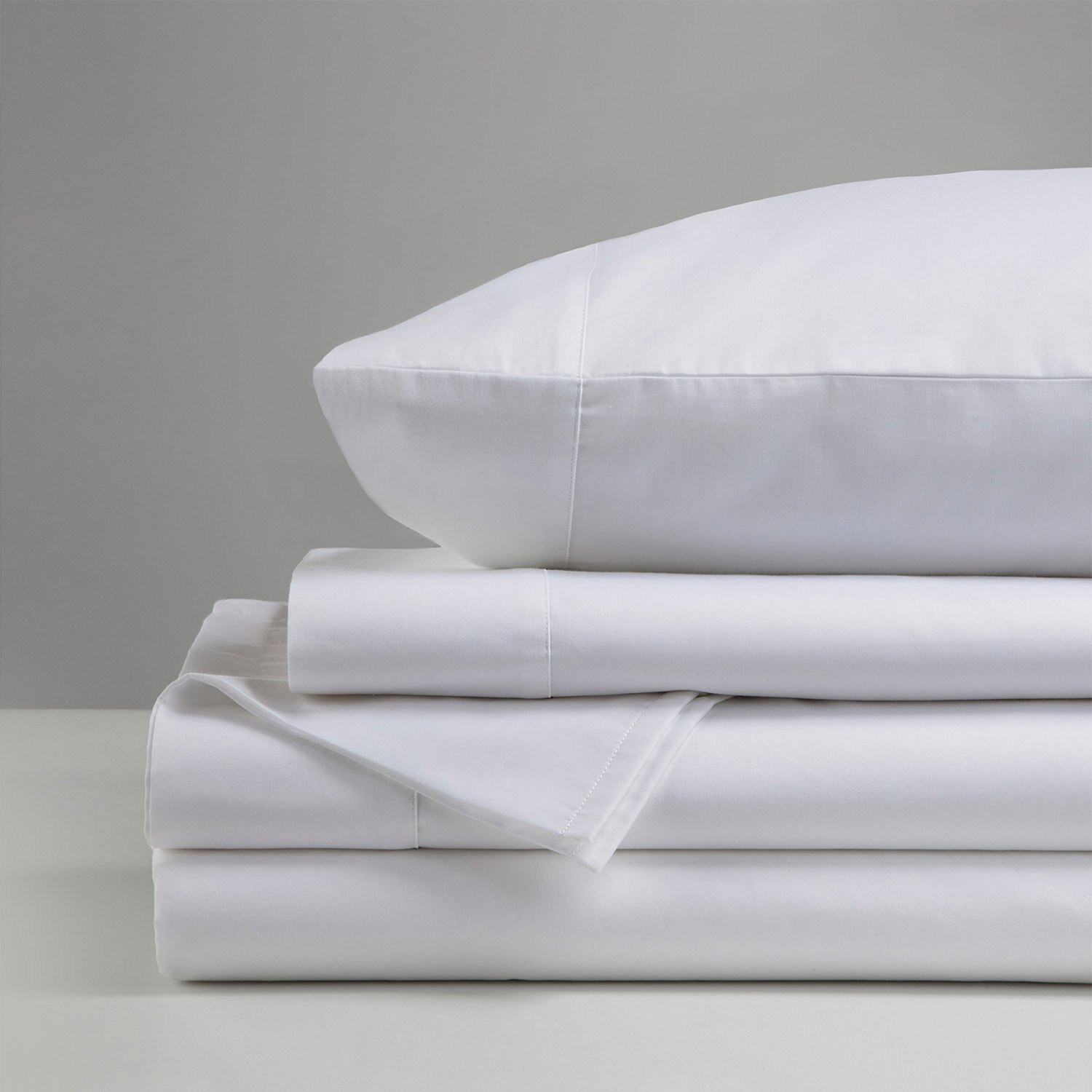 A neatly folded stack of Gryphon Collection - Sheets - Solid by WestPoint Home, topped with a matching pillow, set against a plain light gray background.